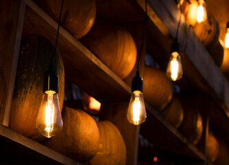 Pumpkins on wooden shelves illuminated by warm glowing vintage bulbs. Cozy rustic autumn atmosphere background for fall, harvest, farmers markets and Halloween themes. Happy Thanksgiving Day.