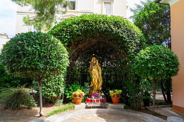 golden statue of the Virgin Mary Madonna placed under a green leafy arch with potted plants and red devotional candles in front. religious shrine or grotto in an outdoor garden in Opatija Croatia