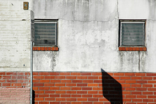 A facade with two plain windows on a weathered white concrete wall with a red brick base, a perfect example of the effects the weather have on the built environment. Good for post about urban decay