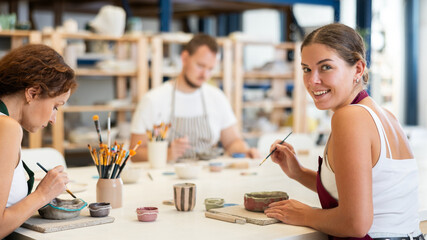 In pottery workshop, two girls and guy are sitting at table, busy coloring products made of raw...