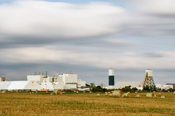 Round hay bales on farmland in Saskatchewan, Canada, with large white industrial buildings and storage domes in the background under dramatic cloudy sky.