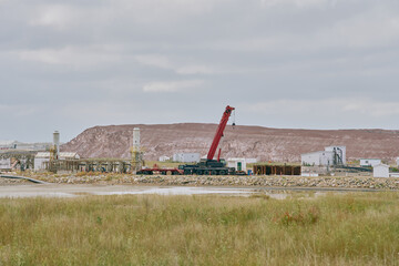 A red crane operates at an industrial facility near a potash mine in Saskatchewan, Canada, with pipelines, storage buildings, tailings pile, and a rocky embankment by the water.