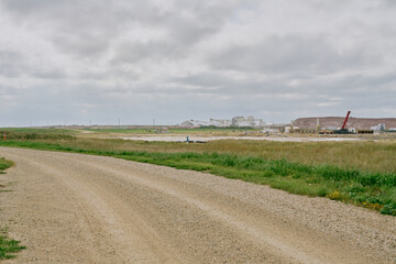 A gravel country road curves through the prairie landscape toward an industrial potash mine in Saskatchewan, Canada, with processing buildings, tailings pile, and cloudy sky in the distance.