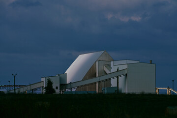 Main processing building of a potash plant in Saskatchewan, Canada, with white industrial architecture, ventilation stacks, and smoke rising under dramatic dark clouds.