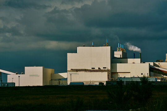 Main processing building of a potash plant in Saskatchewan, Canada, with white industrial architecture, ventilation stacks, and smoke rising under dramatic dark clouds.