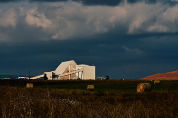 Industrial potash facility in Saskatchewan, Canada, with white processing buildings, conveyor systems, hay bales in the foreground, and tailings pile under dramatic cloudy sky.