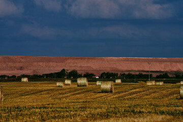 Round hay bales on farmland in Saskatchewan, Canada, with PotashCorp freight train cars and a large potash mine tailings pile in the background under dramatic cloudy sky.