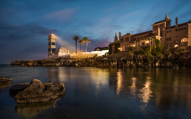 Obraz premium Scenic lighthouse at night with illuminated coastal architecture, palm trees and reflections in calm ocean water Portugal