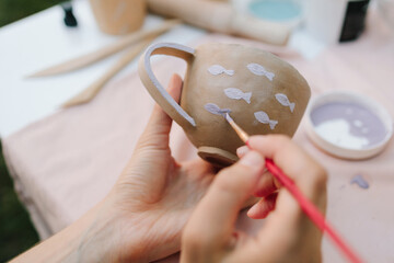 Ceramic painting of mug with fish pattern using brush strokes. Artist paints delicate purple fish on a handmade clay mug. Painting ceramic mug with purple fish using brush and paint.