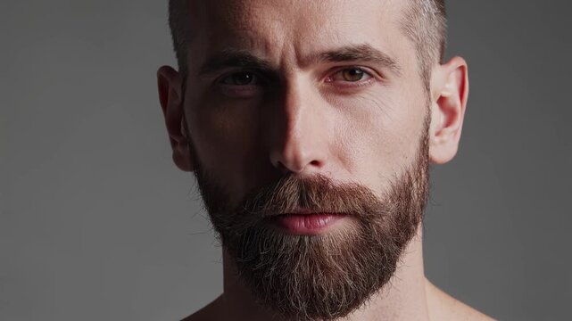 Man with neatly trimmed beard and short hair stands in a neutral studio with gray background emphasizing masculinity minimalism and confident modern style