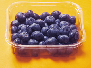 A plastic tray of blueberries sits on a bold orange background. The tray is clear and filled with fresh organic fruits ready for consumption. Healthy diet concept. Natural product.