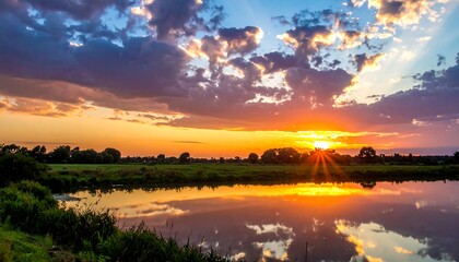 Vivid sunset paints the sky with golden hues, reflecting on still water, over green landscape, with fluffy clouds