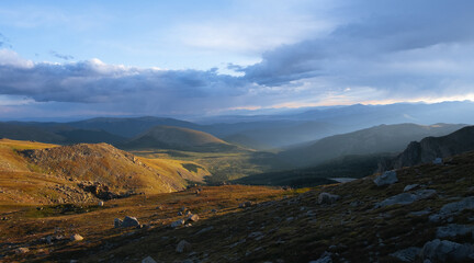 Sun rays breaking through clouds over mountain valley, scenic wilderness landscape with rolling hills and dramatic light