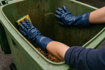 Cleaning The Trash Can: The image features hands in blue gloves scrubbing the interior of a dirty trash bin. A scene emphasizing cleanliness and hygiene.