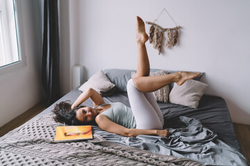 Playful young woman lying on bed with leg raised, smiling at camera, expressing balance between movement, inner freedom and spontaneous joy in personal space.
