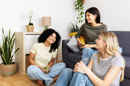 Three young women enjoying each other's company, laughing and sharing a healthy salad while relaxing together in a cozy living room. Female friendship and youth concept