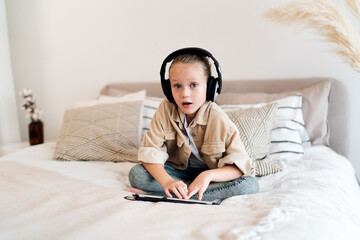 Surprised little girl in headphones looking up from tablet with wide eyes, sitting on bed in cozy...