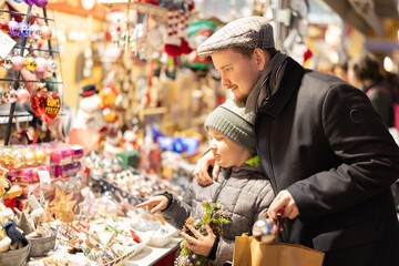 Man with son among crowd of shoppers at holiday fair. Buyers by outdoor souvenir shop window examines Christmas toys and interior decorations, colorful balls, figurines and tinsel..