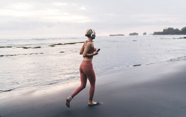 Woman runs barefoot on black sand near waterline at sunrise with smartphone and headphones, embodying tech-powered rhythm, feminine strength and personal audio-driven movement.