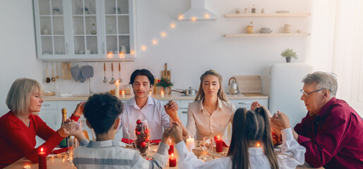 Multi generation family holding hands and praying before Thanksgiving or Christmas dinner, sitting...