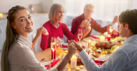 Young woman celebrating Christmas with her husband and senior parents, having festive Xmas dinner, praying to God, holding hands with family at table indoors. Winter holidays at home concept