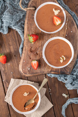 Three white bowls of rich chocolate mousse, one with a golden spoon, are elegantly arranged on a wooden board and table with fresh strawberries and delicate white flowers