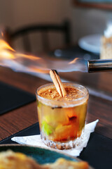 Colorful fruity cocktail with garnish in a medium-sized, round glass. A cinnamon stick and fire.