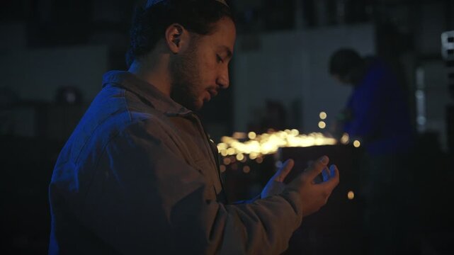 A young man praying during prayer inside a workshop, raising his hands in supplication while sparks fly in the background. A symbolic moment combining faith and daily work life.