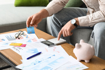 Young man in debt with credit card using calculator at home, closeup