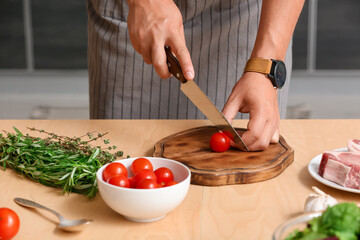 Young man cutting tomato at table in kitchen, closeup