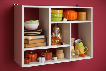 Shelf with different dishes, books and autumn decor hanging on color wall