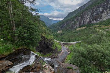 Waterfall cascades into a valley with a bridge and road.