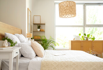 Fototapeta premium Beautiful interior of light bedroom with wooden chest of drawers, window, houseplants and bed