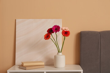 Vase with gerbera flowers, books and picture on shelving unit near beige wall in room