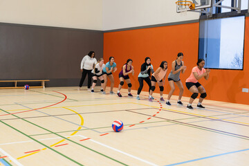 Diverse female teammates crouching on court at gym with volleyball and african american coach