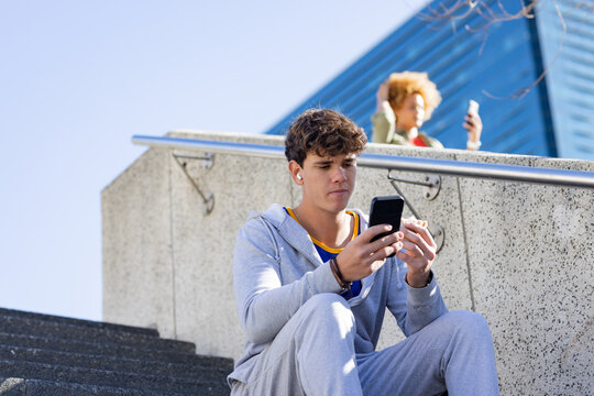 Man sitting on steps with smartphone and earbuds in plaza, woman by railing checking phone