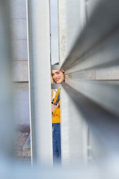 Peeking woman in yellow sweater, jeans carrying backpack through metal beams on urban walkway