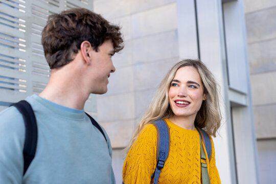 Diverse classmates walking and chatting outside stone facade with backpacks near metal panel
