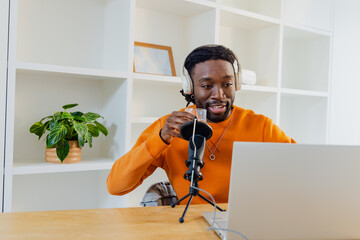 African American man at desk wearing sweater headphones speaking into microphone with mug at laptop