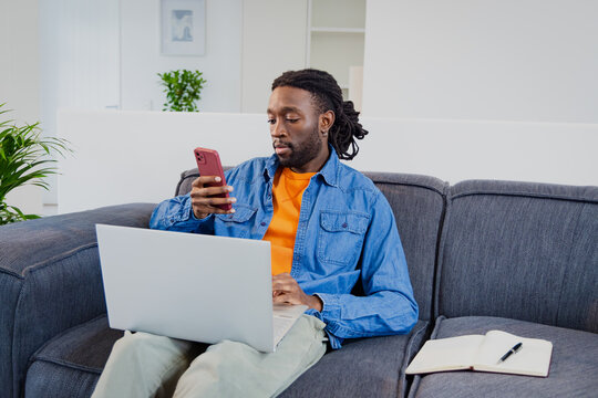 African American man typing on laptop and scrolling smartphone on living room sofa with notebook
