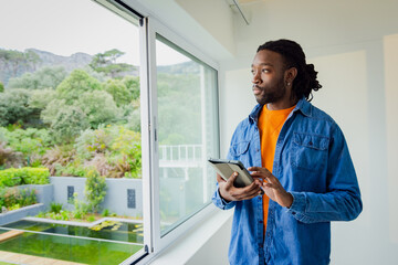 African American man standing beside sliding glass window at home holding tablet and gazing at pond