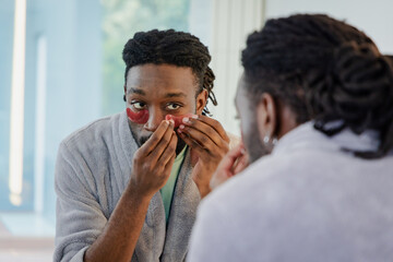 African American man applying red gel eye patches under eyes while standing at bathroom mirror