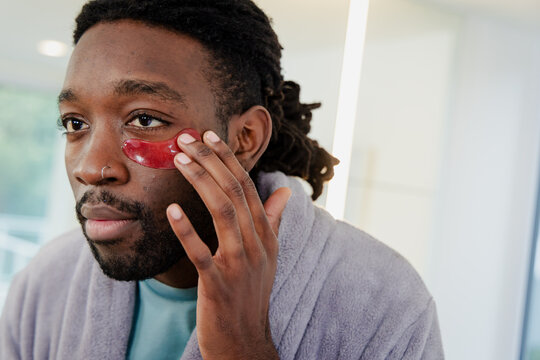 African American man applying red hydrogel under-eye patch wearing bathrobe in bathroom - Powered by Adobe