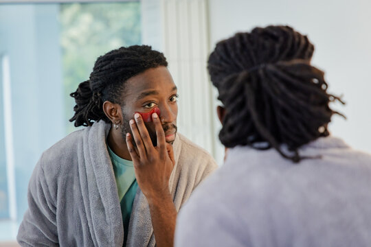 Adult African American man standing at sink by mirror wearing bathrobe applying red under-eye patch