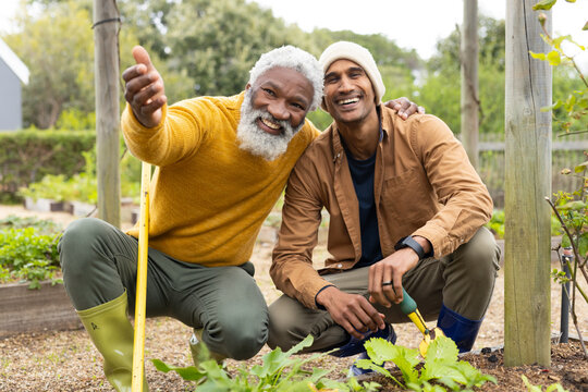 Senior African American friends kneeling in garden holding hoe and trowel by beds