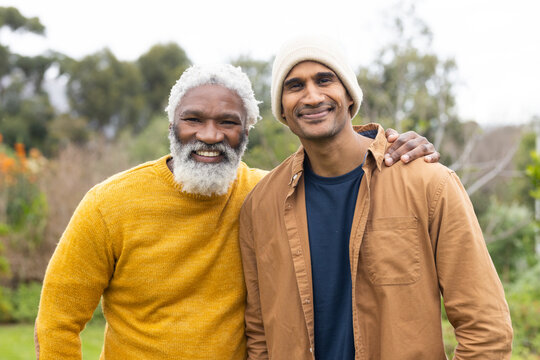 African American senior father and Indian mature son standing in garden with flowering shrubs