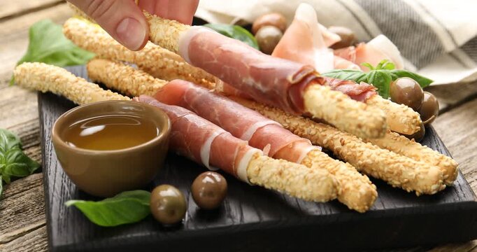 Woman taking grissini breadstick with prosciutto from board at wooden table, closeup