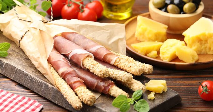 Tasty grissini breadsticks with prosciutto and cheese on wooden table, closeup. Camera moving right