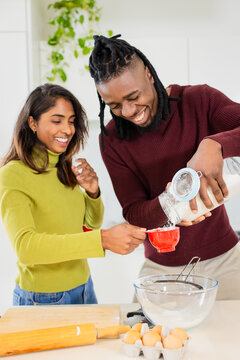 Diverse couple wearing green turtleneck baking at kitchen counter using red measuring cup flour jar