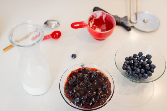 Blueberry compote bowl sitting on counter with milk carafe, blueberries and baking tools